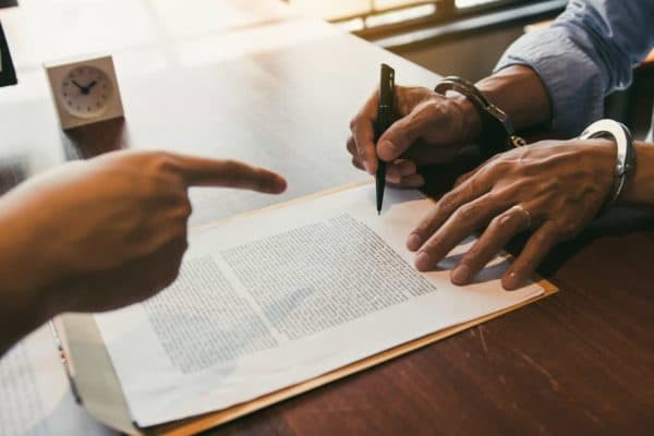 A handcuffed man signs documents while someone points at him.